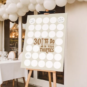 May include: A beige sign on a wooden easel with the text "30 Things To Do In Your Thirties" in brown. The sign has white circles for checklist items. White balloons are in the background.