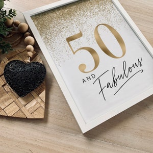 May include: A white framed print featuring the gold text 50 AND FABULOUS with gold glitter accents at the top. Beside the frame is a black metal heart ornament attached to a string of natural wooden beads and a small green plant sprig.