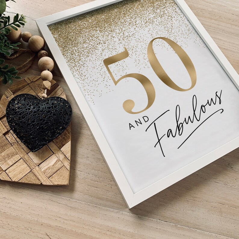 May include: A white framed print featuring the gold text 50 AND FABULOUS with gold glitter accents at the top. Beside the frame is a black metal heart ornament attached to a string of natural wooden beads and a small green plant sprig.