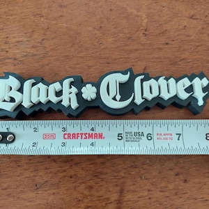 May include: A black and white "Black Clover" logo, approximately 7 inches long, is displayed on a wooden surface. The logo is measured with a Craftsman tape measure.