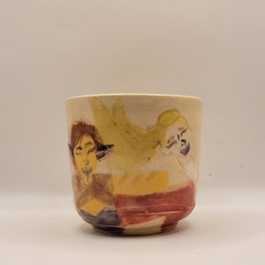 May include: A handmade ceramic cup with a beige exterior. The cup features a painted design of two figures in yellow, red, and brown. The cup has a rounded base and a slightly flared rim.