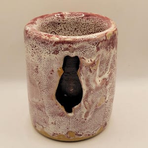 May include: A handmade ceramic cup with a unique design. The cup is cylindrical, with a speckled white and burgundy glaze. A dark, irregular opening is cut into the side. The rim and base have a light brown color.