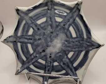 May include: A ceramic plate with a unique starburst design. The plate features a white base with a navy blue pattern of concentric circles and radiating lines. The plate is displayed on a metal stand.