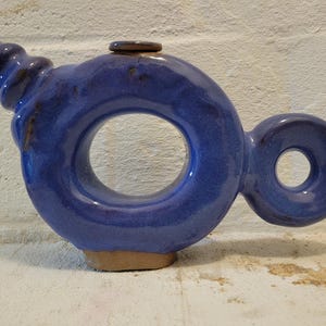 May include: A unique, handmade ceramic teapot in a vibrant blue glaze. The teapot features a large circular opening in the center, a smaller circular handle, and a spiraled spout. The base is a contrasting tan color.