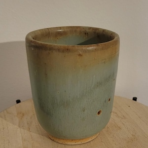 May include: A handmade ceramic tumbler with a cylindrical shape. The cup features a gradient of colors, transitioning from a light brown rim to a pale blue-green body. Small, dark brown spots dot the surface. The base is a lighter brown.