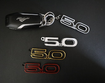 Puede incluir: Cuatro llaveros con el número "5.0" en diferentes colores: blanco, dorado, rojo y negro. Los llaveros están sobre una superficie negra. El llavero superior está unido a una llave de coche negra y plateada con un logotipo de Ford Mustang.
