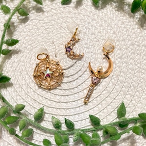 May include: Three gold-colored celestial-themed charms. One is a compass design with a star, another is a crescent moon with a purple star, and the third is a wand with a pearl and pink gem.