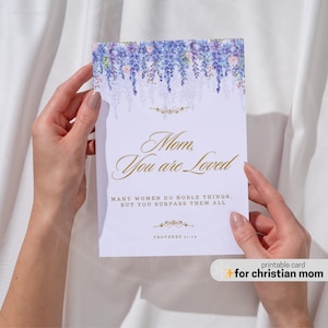 May include: A white greeting card with the text "Mom, You are Loved" in gold script, and a floral design in shades of purple and blue. The card also includes the text "MANY WOMEN DO NOBLE THINGS. BUT YOU SURPASS THEM ALL" and "PROVERBS 31:29".