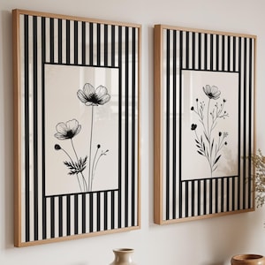 Può includere: Due quadri rettangolari con cornici in legno chiaro caratterizzati da strisce verticali bianche e nere che circondano un pannello centrale bianco con un disegno a linee nere di delicati fiori di campo, disposti fianco a fianco.
