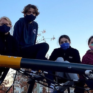 May include: Four people wearing face masks are sitting on a playground structure. The structure is made of yellow and black metal and has a net-like design. The people are sitting on the structure and looking at the camera. The sky is blue and there are trees in the background.