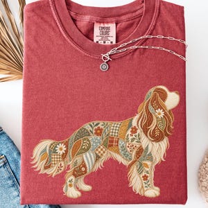 May include: A brick red crewneck t-shirt featuring a graphic of a dog with a patchwork-style coat pattern. The dog illustration incorporates floral, plaid, and leaf motifs in muted earth tones. The shirt has a Comfort Colors brand label at the neck.