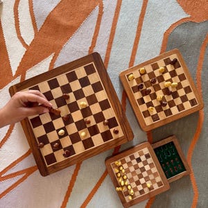 Puede incluir: Tres juegos de ajedrez de madera de diferentes tamaños. El juego más grande está en juego con piezas marrones y amarillas. El juego más pequeño está cerrado con un interior de fieltro verde.