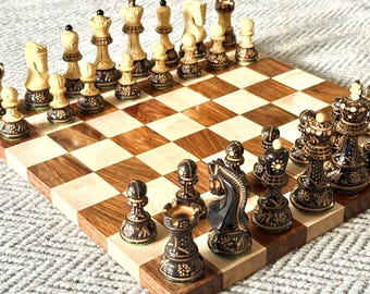 Può includere: Un set di scacchi in legno con una scacchiera a scacchi marrone chiaro e scuro. I pezzi degli scacchi sono intagliati in modo intricato in una combinazione di colori marrone chiaro e scuro a contrasto. I pezzi sono disposti sulla scacchiera, pronti per giocare.