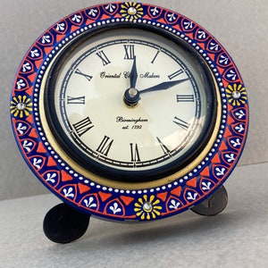 May include: A blue and red clock with a white face and black hands. The clock has a decorative border with yellow flowers and rhinestones. The clock face reads "Oriental Clock Makers Birmingham est. 1792".