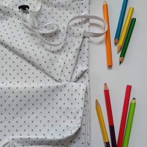 Puede incluir: Una bolsa de cordón blanco con lunares grises y un cordón blanco. La bolsa está doblada y colocada sobre una superficie blanca. Hay ocho lápices de colores al lado de la bolsa.