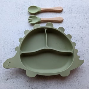 May include: A sage green dinosaur-shaped divided plate with a matching spoon and fork set. The plate has three compartments and a smiling face. The spoon and fork have wooden handles and green silicone heads. A fun and functional set for mealtime.