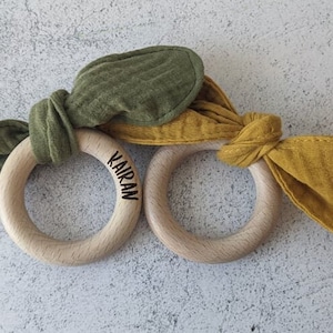 Könnte beinhalten: Zwei Baby-Beißringe aus Holz mit Stoff-Hasenohren in Olivgrün und Senfgelb. Ein Ring hat den Namen "KAIRAN" eingraviert. Die Ringe liegen auf einer hellgrauen Oberfläche.