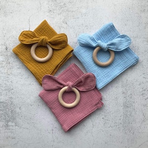 May include: Three square muslin cloths in yellow, blue, and pink with wooden rings tied in the center to create a bow.