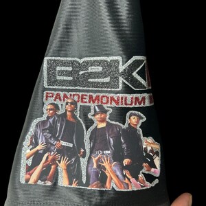 May include: Black bucket hat with a silver glitter E2K logo and the word "Pandemonium" in red. The hat has a graphic of four people with hands reaching up. The hat is held by a hand.