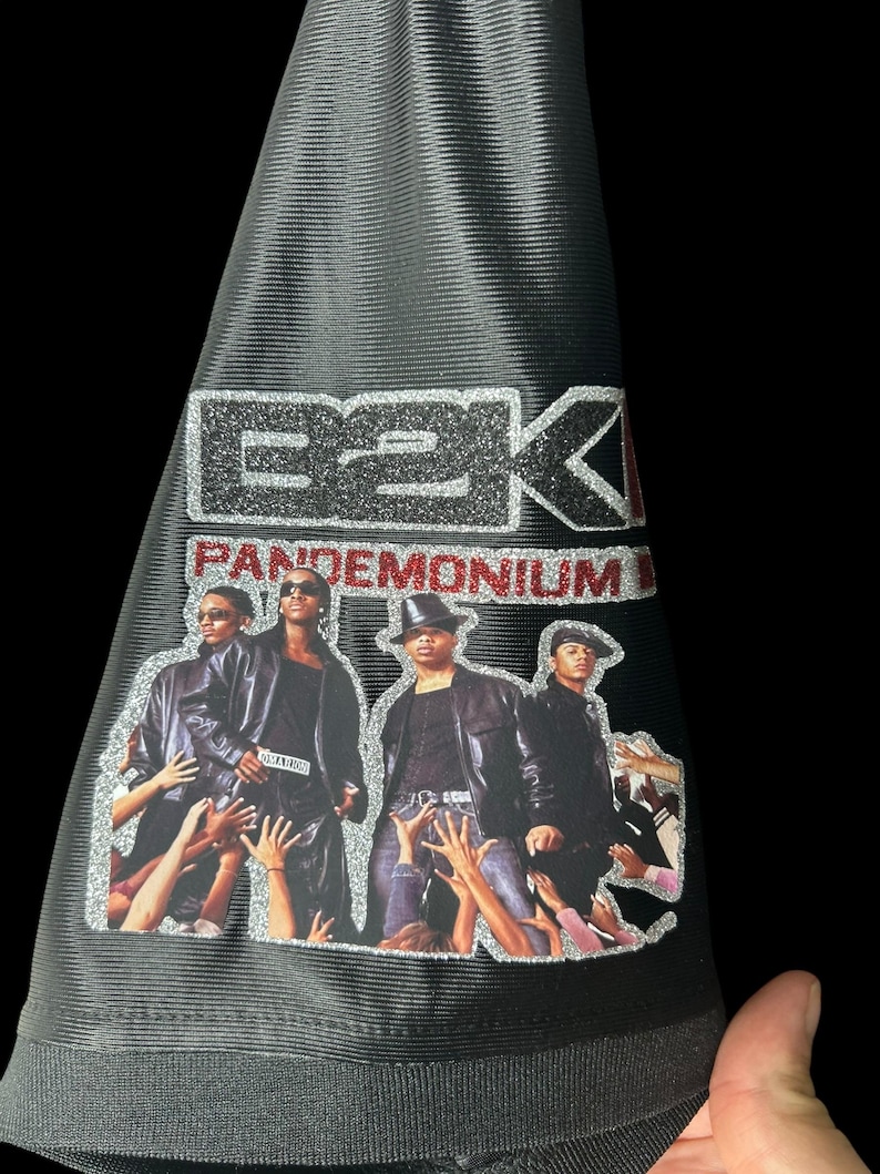May include: Black bucket hat with a silver glitter E2K logo and the word "Pandemonium" in red. The hat has a graphic of four people with hands reaching up. The hat is held by a hand.