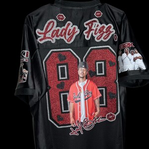 May include: Black jersey with the name "Lady Fizz" in red glitter script. The number "88" is in red glitter with a photo of a person in a red baseball jersey. The jersey has a black mesh design and features lip graphics.