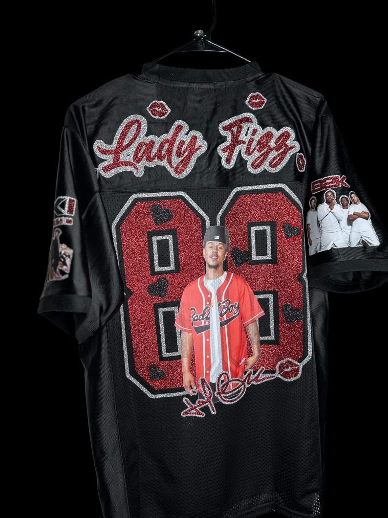May include: Black jersey with the name "Lady Fizz" in red glitter script. The number "88" is in red glitter with a photo of a person in a red baseball jersey. The jersey has a black mesh design and features lip graphics.