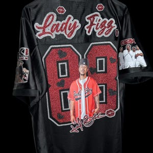 May include: Black jersey with red glitter detailing, displaying the name "Lady Fizz" and the number "88". The jersey features a photo of a person and a signature. The sleeves have images of people.