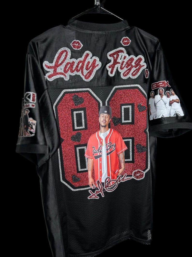 May include: Black jersey with red glitter detailing, displaying the name "Lady Fizz" and the number "88". The jersey features a photo of a person and a signature. The sleeves have images of people.