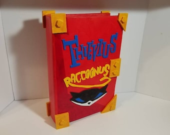 Peut inclure: Un livre rouge avec des accents dorés et le titre "Thievius Raccoonus" en lettres bleues et jaunes. Le livre présente un chat noir avec des yeux bleus et un nez rouge.