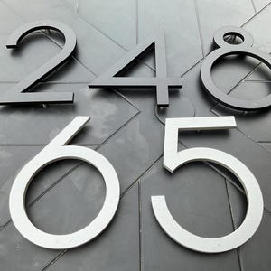 May include: A selection of contemporary house numbers in different sizes and fonts. The numbers include a black 2, 4, and 8, and silver 6 and 5. The numbers are displayed on a dark grey, textured surface.