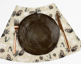 May include: A brown ceramic plate with a white linen placemat featuring a pine cone pattern. A gold fork and knife are placed on the placemat.
