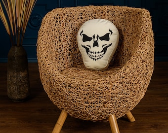 May include: A wicker chair with a skull-shaped pillow. The chair has a woven design and three wooden legs. The pillow is white with a black skull design.