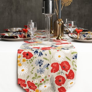 Op de afbeelding: Een beige tafelkleed met een bloemenpatroon met rode klaprozen, witte madeliefjes en blauwe wilde bloemen. De loper ligt op een wit tafelkleed met twee couverts en een middelpunt van zwarte kaarsen in glazen houders.