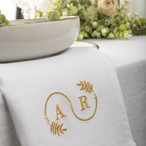 May include: White linen napkin with gold embroidery of the letters "A" and "R" in a circle with a leafy design.