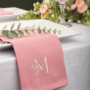 Könnte beinhalten: Eine rosa Leinen-Serviette mit einem weißen, gestickten Blumenmonogramm "M" auf einer weißen Tischdecke mit einem weißen Teller und einem Glas rosa Getränk.