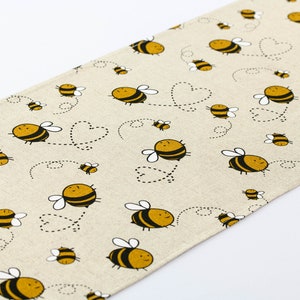 May include: A beige table runner with a repeating pattern of yellow and black bees flying with hearts. The bees have white wings and black eyes.