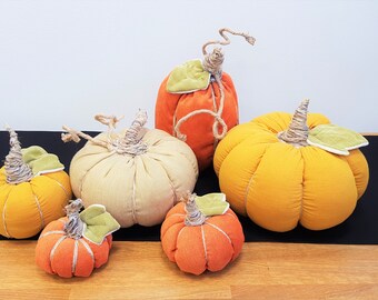 May include: Five fabric pumpkins in various sizes and colors. The pumpkins are orange, yellow, and beige. They are all sewn with a simple design and have a stem made of twine.