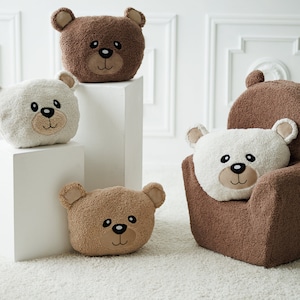 Op de afbeelding: Vier pluche teddybeer-vormige kussens in bruin en wit. Twee kussens staan op witte vierkante sokkels, één zit in een bruine pluche fauteuil en één ligt op de vloer. Alle kussens hebben geborduurde gezichten met zwarte ogen en neuzen.