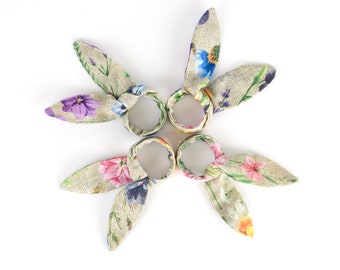 Puede incluir: Cuatro anillos de servilleta de tela floral en forma de orejas de conejo. Los anillos son beige con un patrón floral multicolor.