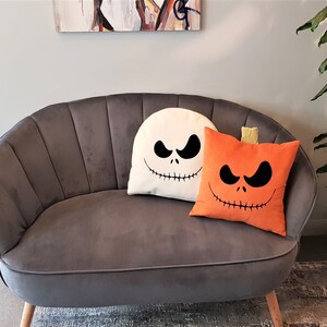 May include: A gray velvet sofa with two decorative pillows. One pillow is white with a black Jack Skellington face and the other is orange with a black pumpkin face.