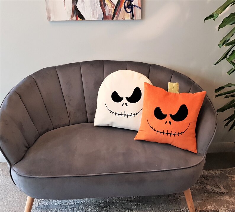 May include: A gray velvet sofa with two decorative pillows. One pillow is white with a black Jack Skellington face and the other is orange with a black pumpkin face.