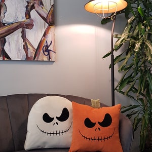 May include: Two Halloween themed throw pillows on a gray couch. One pillow is white with a black Jack Skellington face. The other pillow is orange with a black pumpkin face.