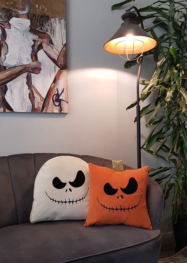 May include: Two Halloween themed throw pillows on a gray couch. One pillow is white with a black Jack Skellington face. The other pillow is orange with a black pumpkin face.