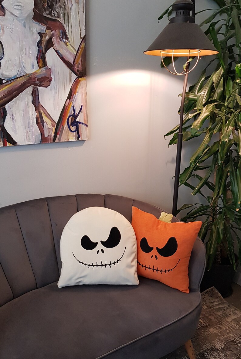 May include: Two Halloween-themed throw pillows on a gray couch. One pillow is white with a black Jack Skellington face, and the other is orange with a black pumpkin face.