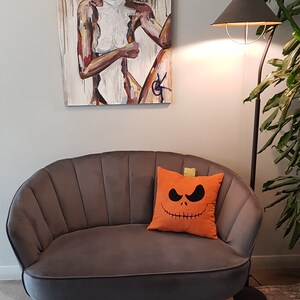 May include: A gray velvet tufted sofa with a decorative orange pillow featuring a black Jack Skellington pumpkin face. The sofa is in front of a white wall with a painting of a woman in the background.