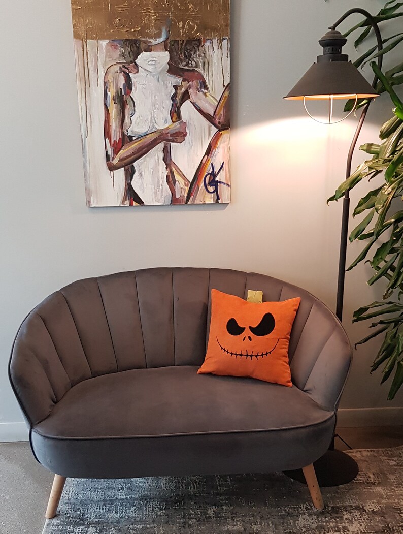 May include: A gray velvet tufted sofa with a decorative orange pillow featuring a black Jack Skellington pumpkin face. The sofa is in front of a white wall with a painting of a woman in the background.
