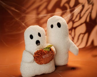 May include: Two white plush ghost figurines, one holding a small orange pumpkin with the word "Hey Boo" embroidered on it. The ghosts are on a brown background with a spiderweb pattern.