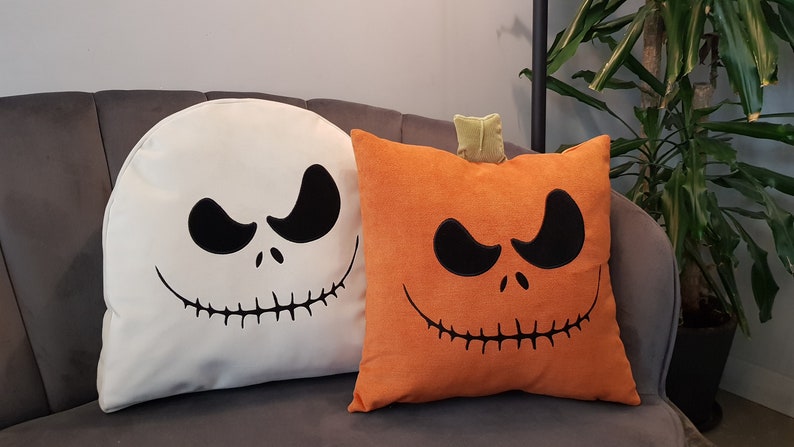 May include: Two Halloween themed throw pillows. One is white with a black Jack Skellington face and the other is orange with a black pumpkin face.