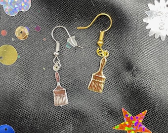 May include: A pair of artisan earrings featuring a silver and gold paint brush charm. The earrings are displayed on a black surface with colorful star and flower-shaped sequins.