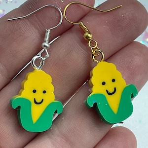 Può includere: Un paio di orecchini con un ciondolo a forma di pannocchia di mais sorridente. Un orecchino ha un gancio d'argento e l'altro un gancio d'oro.